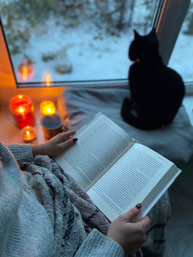 a woman reading a book in front of a window with candles on the windowsill