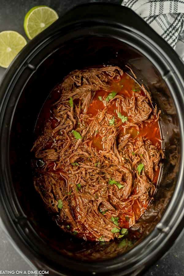 shredded meat in a slow cooker with garnish and limes on the side