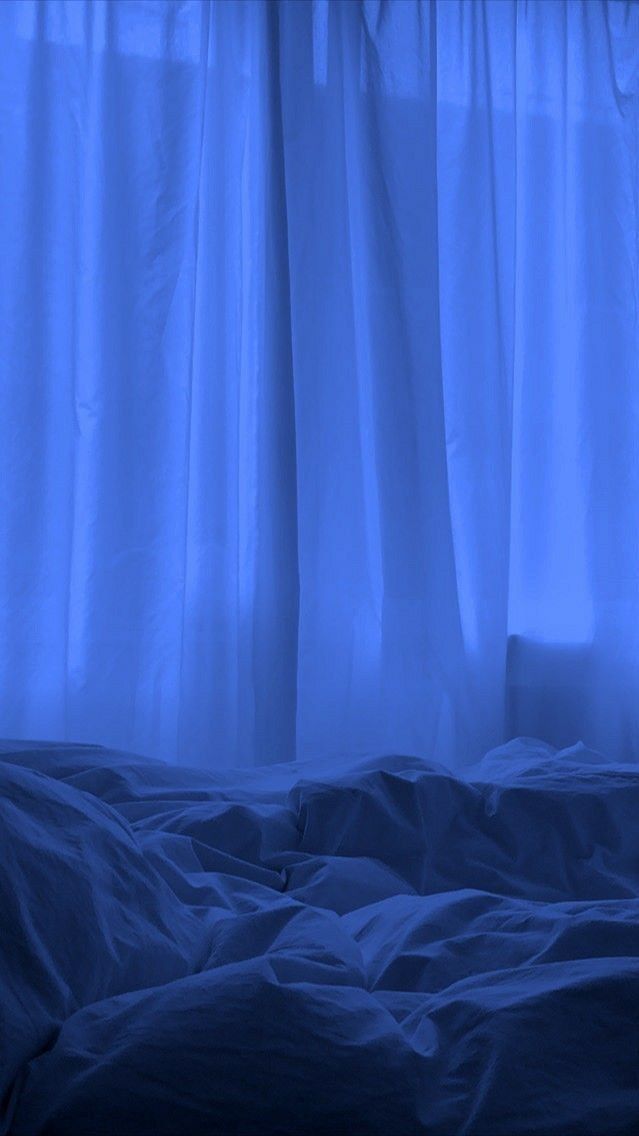 an unmade bed in front of a window with blue light coming through the curtains