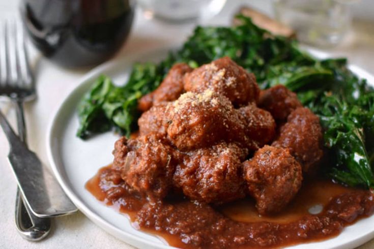 a white plate topped with meatballs covered in sauce and spinach next to a glass of wine