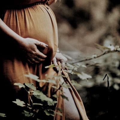 a pregnant woman standing in the woods with her hands on her belly and wearing a brown dress