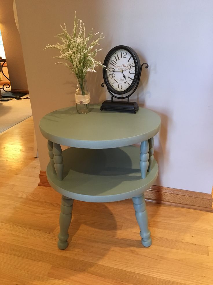 a small green table with a clock on top and flowers in a vase next to it