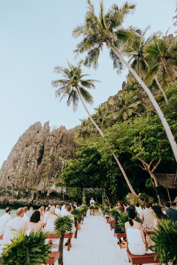 a wedding ceremony on the beach with palm trees and people sitting in chairs looking out at the water