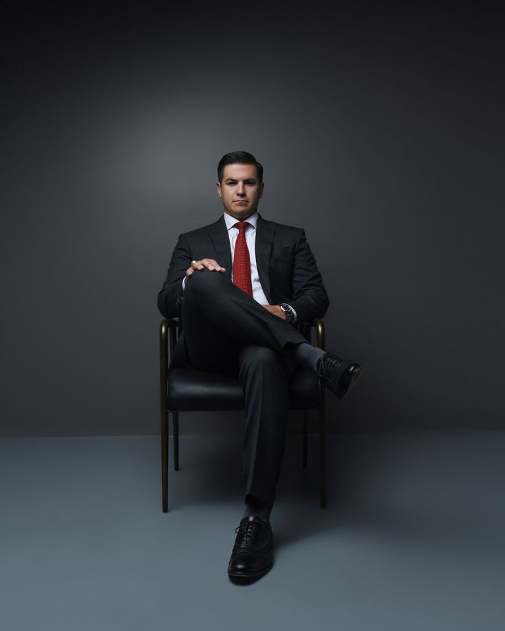 a man in a suit and tie sitting on a chair with his legs crossed, looking at the camera