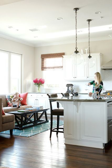 a woman standing in a kitchen next to a living room and dining room table with chairs