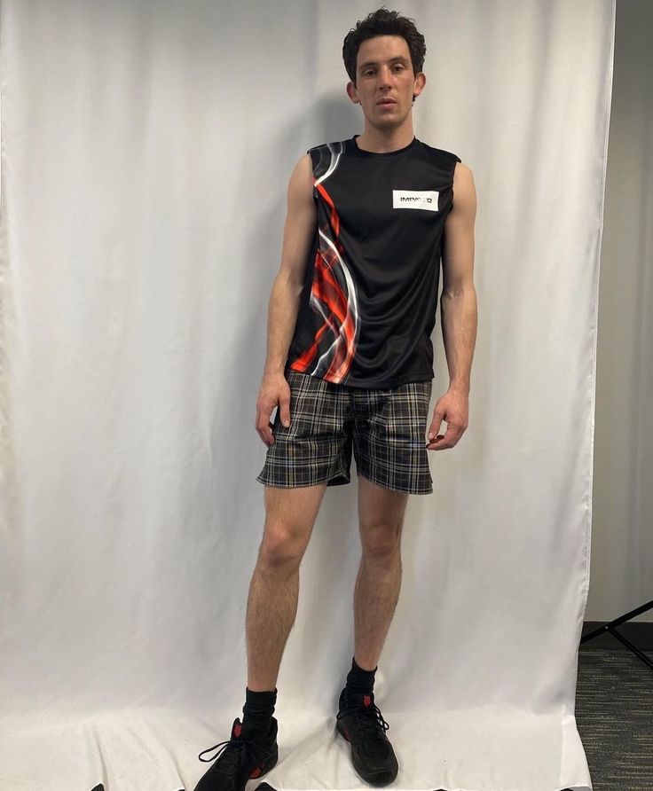 a man standing in front of a white backdrop wearing plaid shorts and a black t - shirt