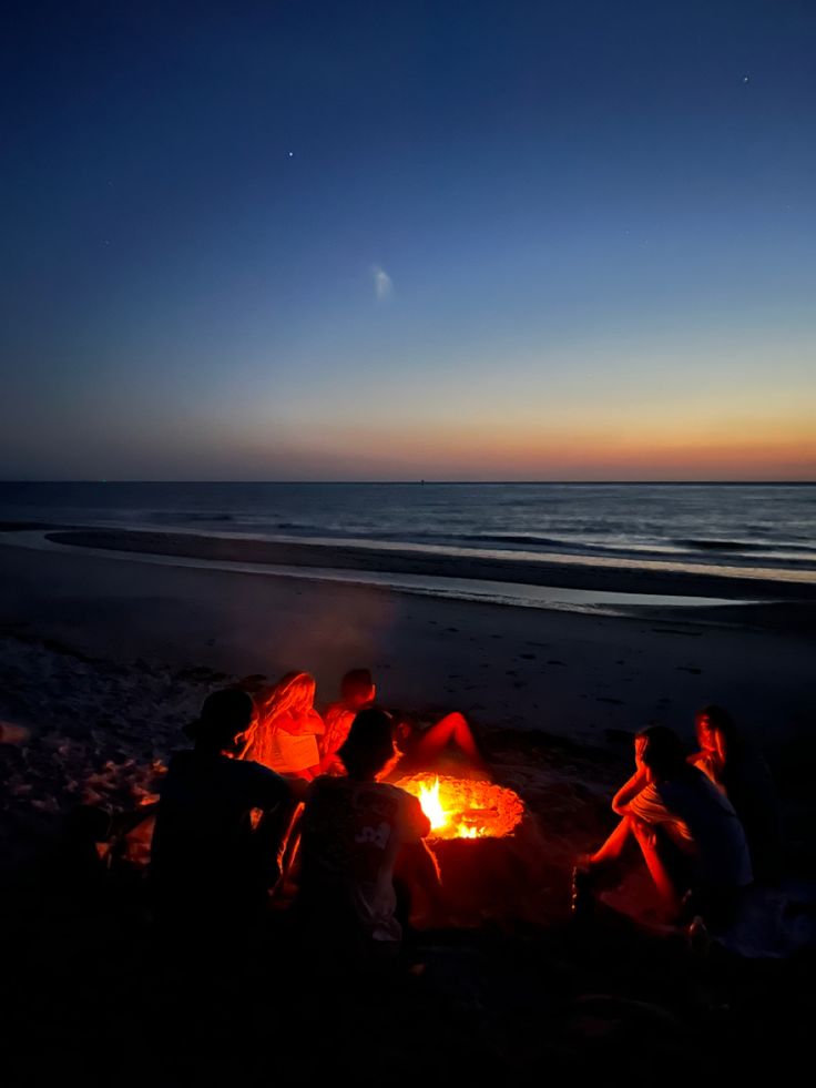 people sitting around a campfire on the beach at night with an ocean in the background