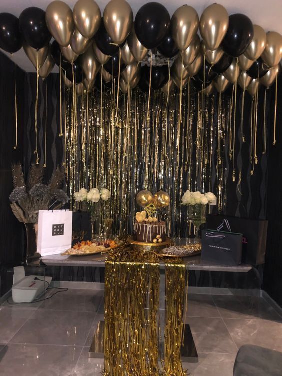 gold and black balloons are hanging from the ceiling above a table with a cake on it