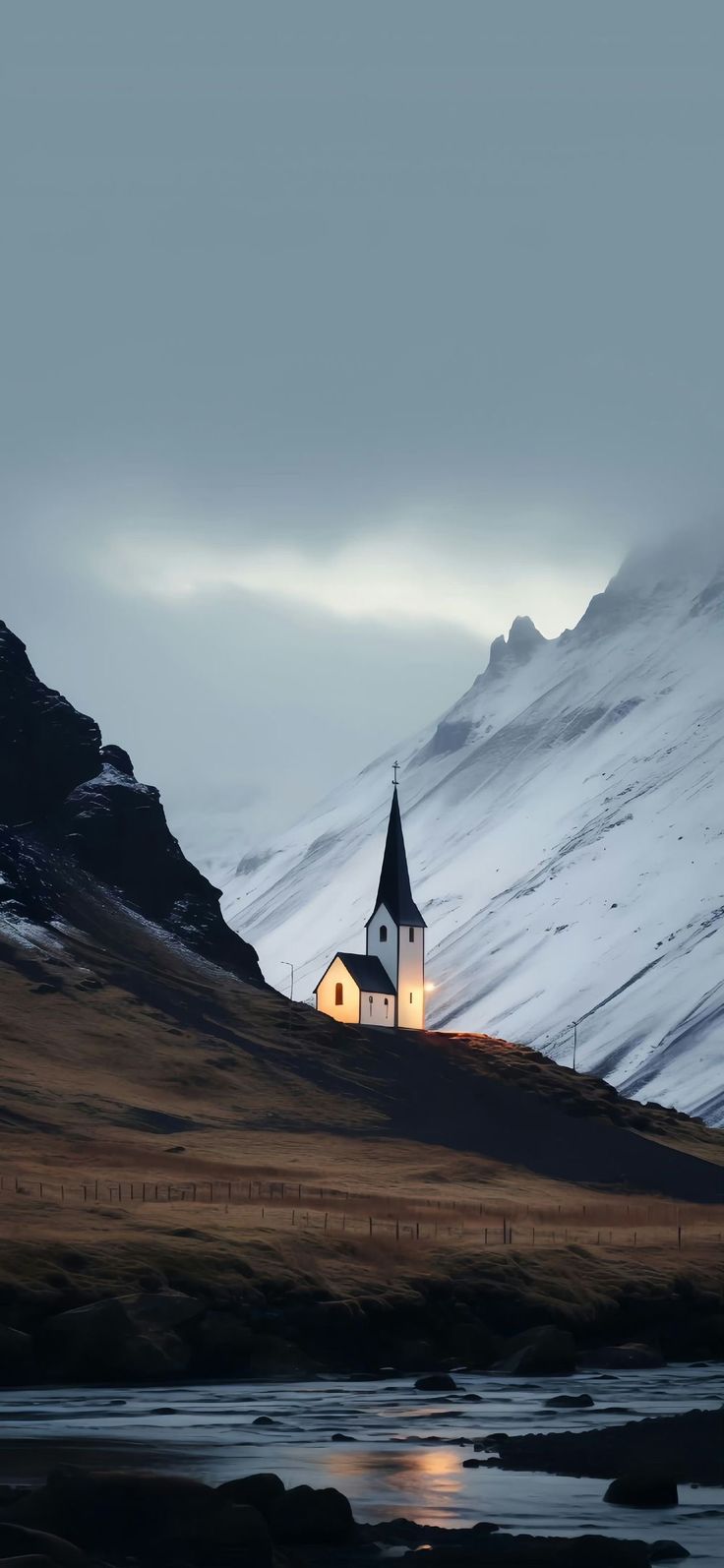 a small church on top of a snowy hill next to a body of water with mountains in the background