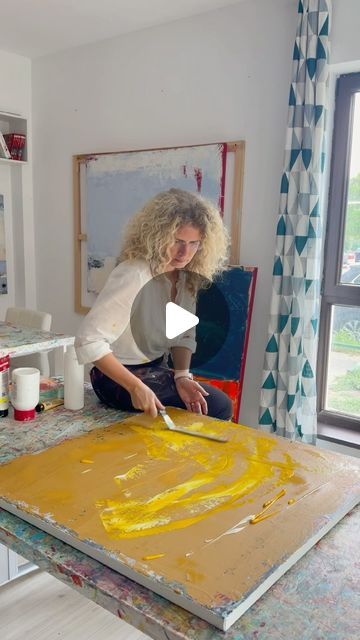 a woman sitting at a table in front of a painting on the easel with yellow paint