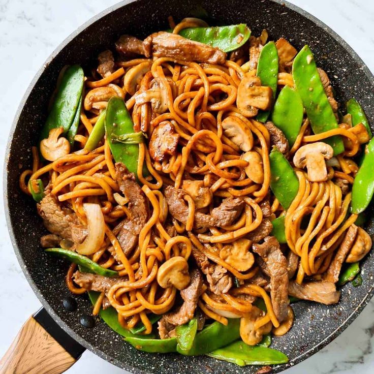 a wok filled with noodles, meat and vegetables on top of a marble counter