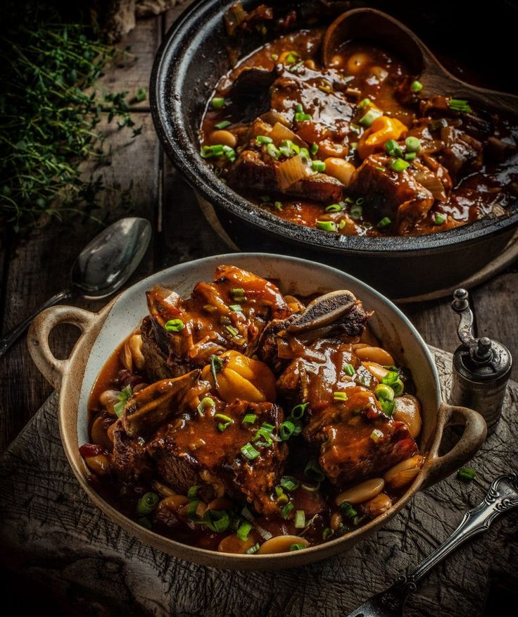 two pans filled with meat and vegetables on top of a wooden table next to spoons