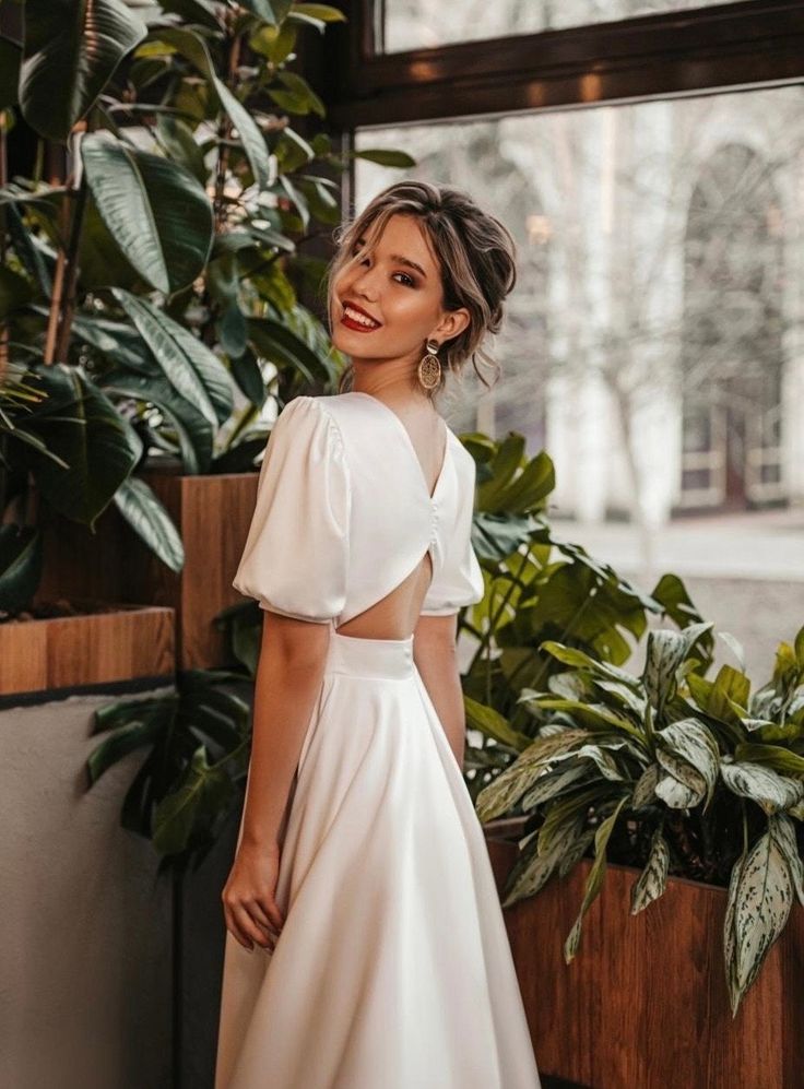 a woman standing in front of a window wearing a white dress