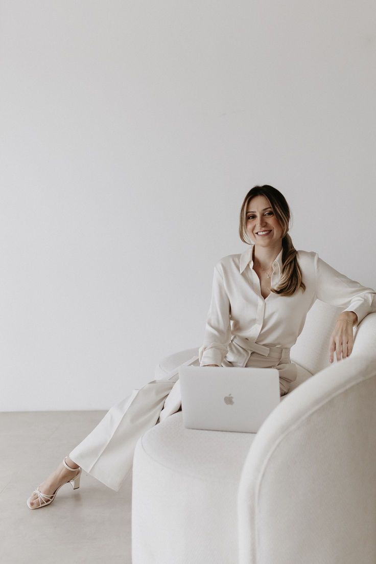 a woman sitting on a white chair with a laptop in her lap and smiling at the camera