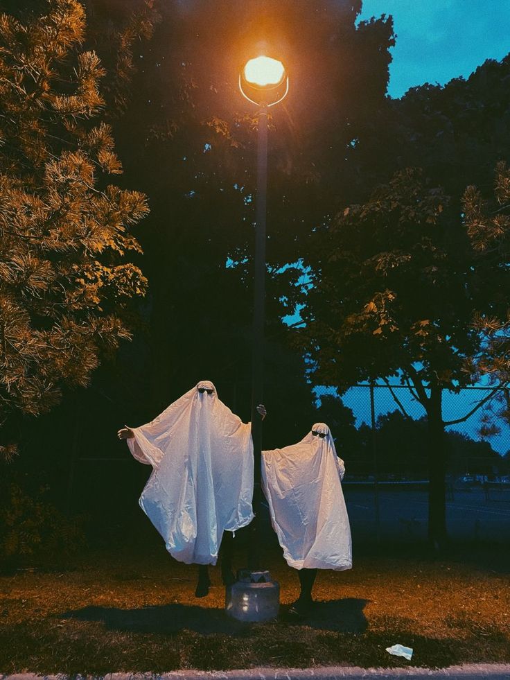 two people in white cloths standing under a street light with their backs turned to the camera