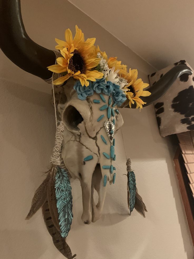 a cow skull with sunflowers and feathers on it's head hanging from the wall