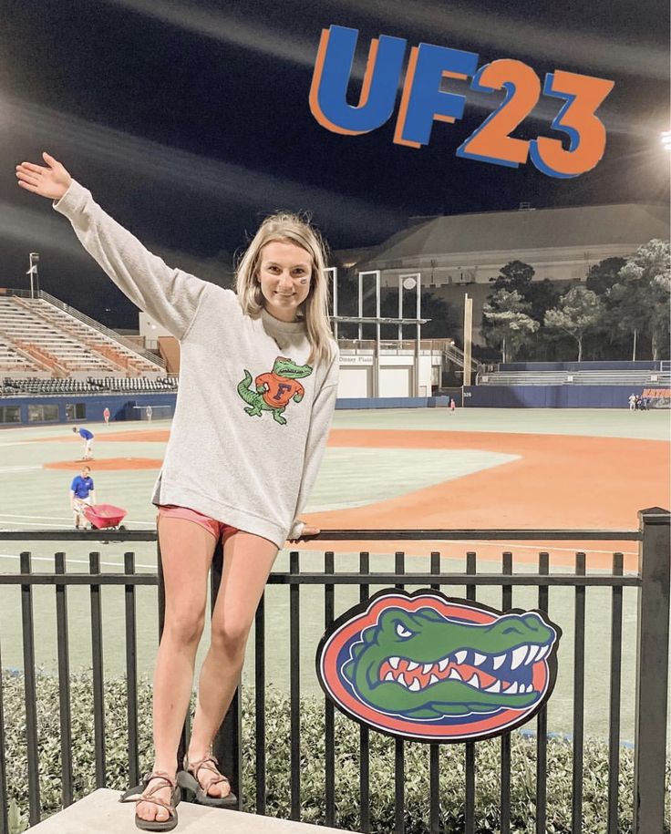 a woman standing on top of a fence next to a baseball field with the uf23 logo