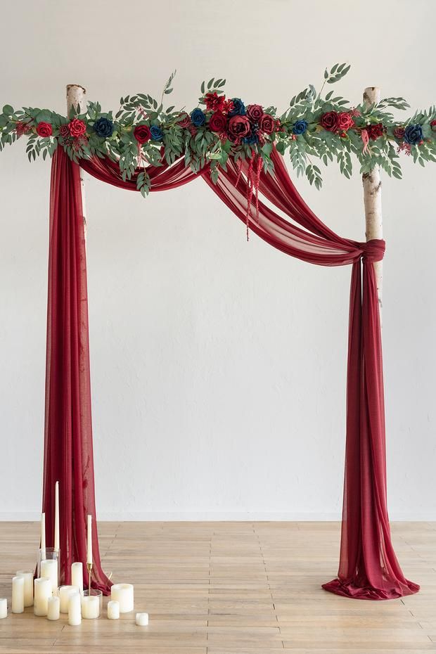 an arrangement of flowers and greenery on a red drape with candles in the foreground