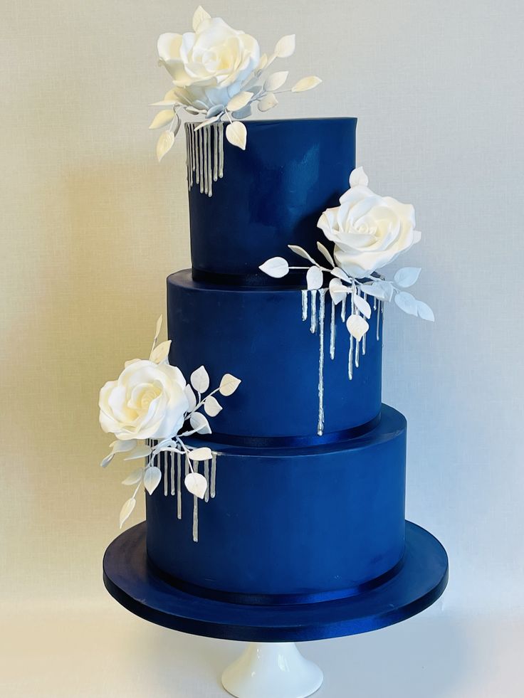 a three tiered blue cake with white flowers on the side and icing dripping from it