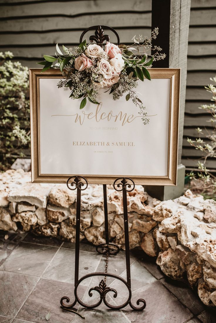 a welcome sign sitting on top of a metal stand next to a stone wall with flowers