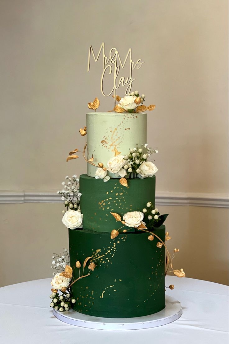 a green wedding cake with white flowers and gold lettering on top is sitting on a table