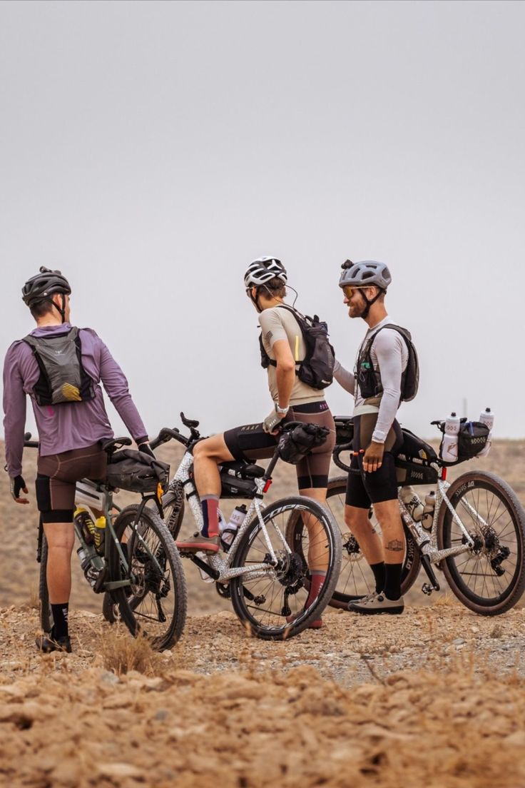 three men standing next to each other with their bikes on the side of a dirt road