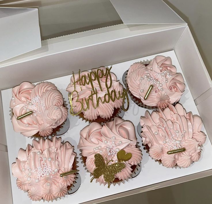 cupcakes with pink frosting and gold decorations are in a white box that says happy birthday