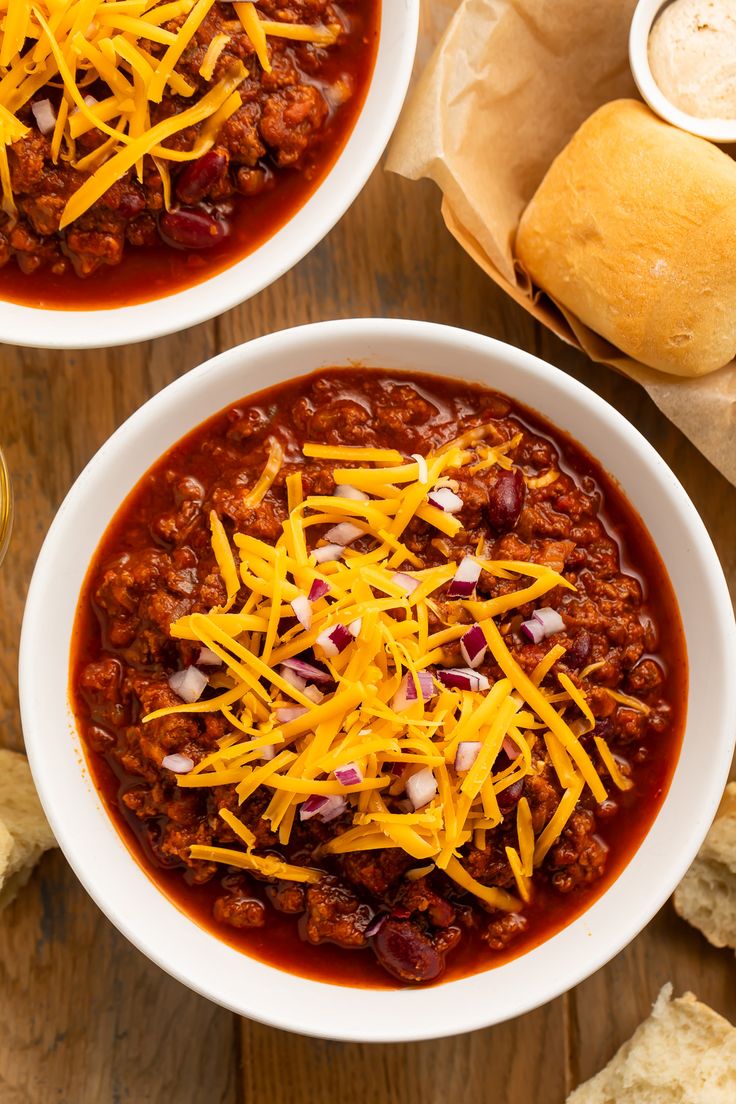 two white bowls filled with chili and cheese on top of a wooden table next to bread