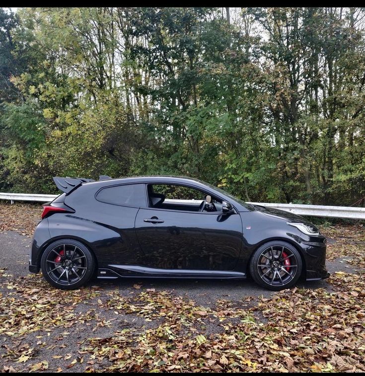 a black car parked on the side of a road in front of trees and leaves