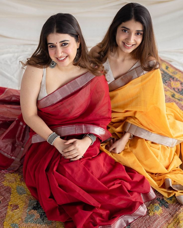 two women in sari sitting on a bed smiling at the camera with their arms around each other