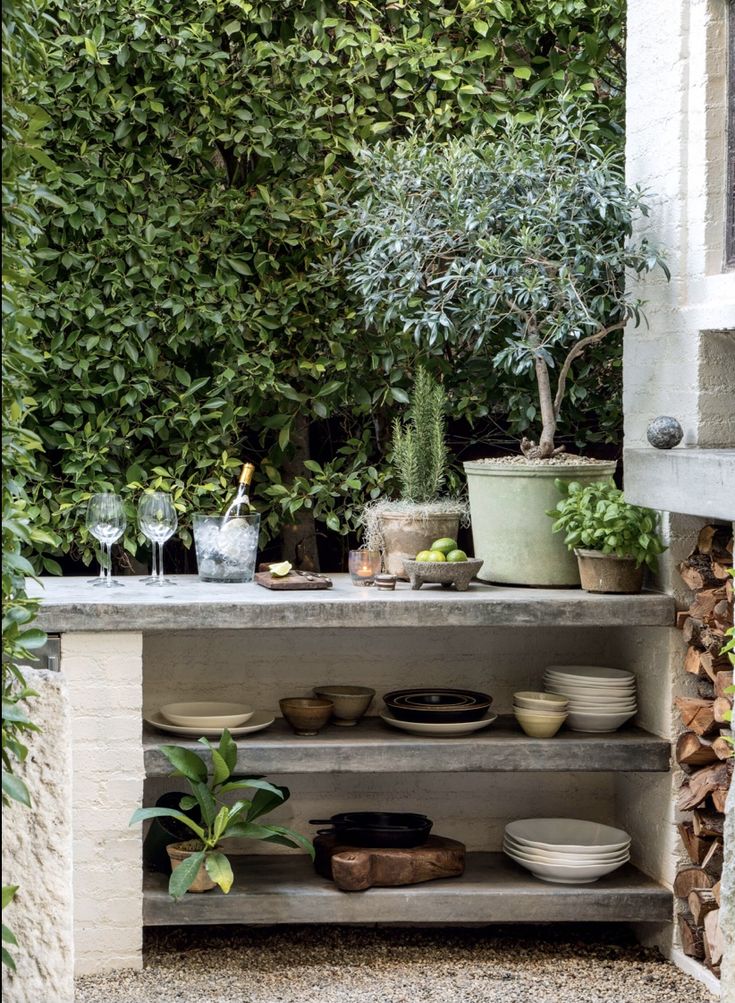 an outdoor kitchen with pots and plates on the counter, potted trees in the background