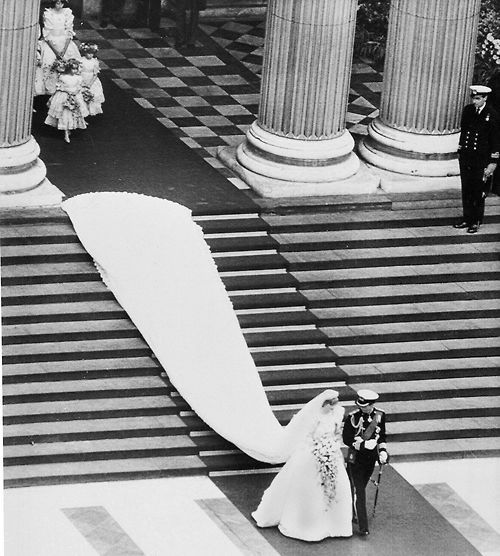 the bride and groom are walking down the aisle in front of columns, dressed in black and white