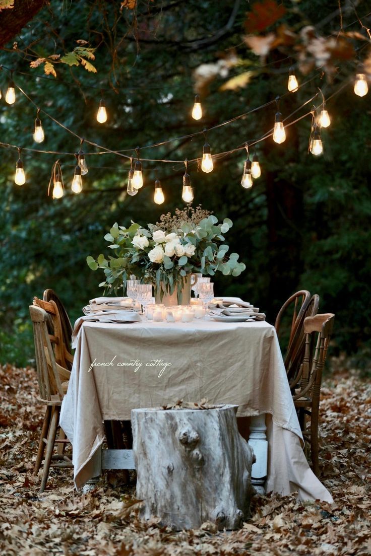 an outdoor dinner table is set with candles and flowers on it, surrounded by leaves