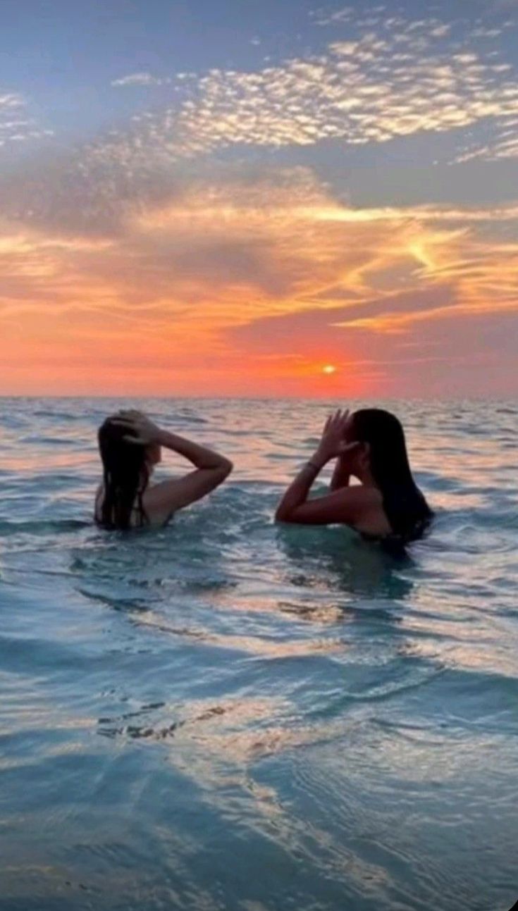 two women in the water at sunset with their backs to each other, one laying on her head