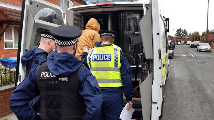 two police officers standing next to the back of a van with its door open and one officer in blue