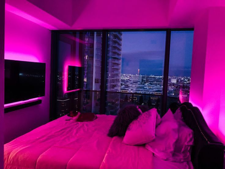 a bedroom with pink lighting and a large window overlooking the cityscape at night