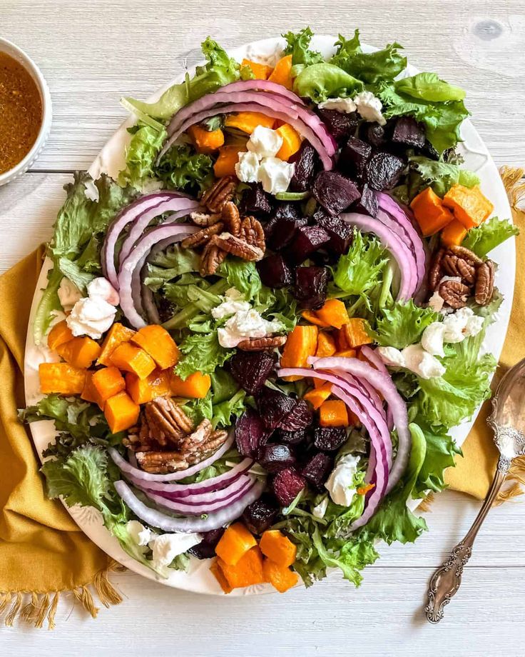 a salad with beets, carrots and pecans on a plate next to a bowl of dressing