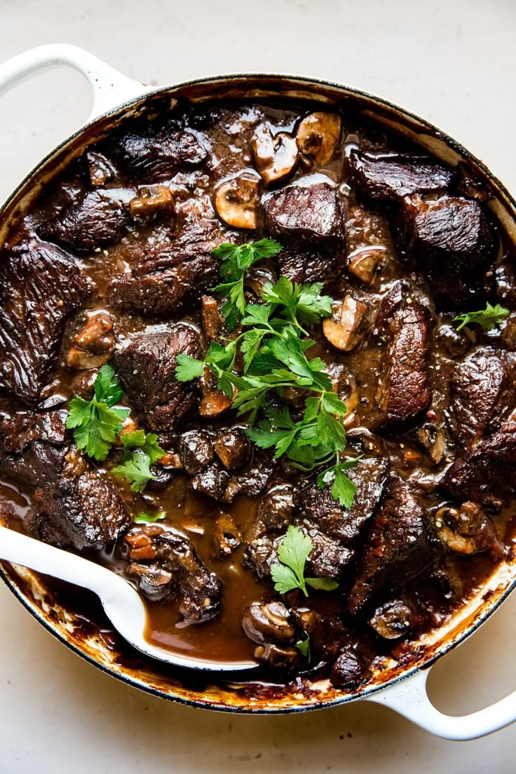 a pot filled with meat and mushrooms on top of a white countertop next to a spoon