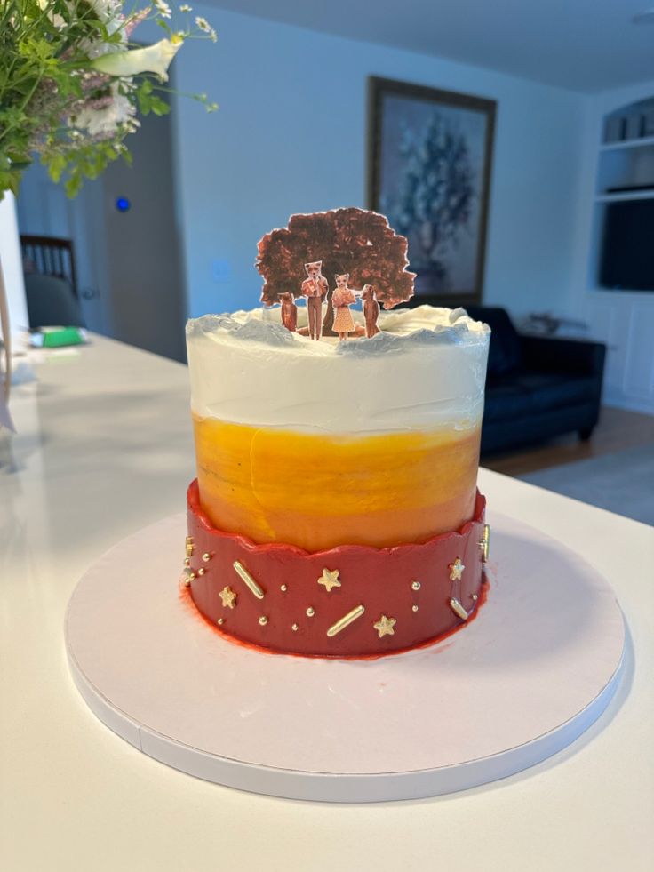 Photo shows a cake sitting on a white counter. The cake is one tier decorated with inspiration from the movie Fantastic Mr Fox. The colors are on the cake are red, orange, and white. The cake is topped with paper cut outs of the characters (Mr Fox, Mrs Fox, Ash, and Christopherson) and their tree home. 
The colors on the cake are in a gradient with red on the bottom, orange in the center, and white on top. The red section has gold star, dots, and bar sprinkles. Fantastic Mr Fox Birthday Party, Wes Anderson Cake, Fantastic Mr Fox Party, Fantastic Mr Fox Aesthetic, Fantastic Mr Fox Movie, Fox Food, Bad Cakes, Fox Cake, Movie Cakes