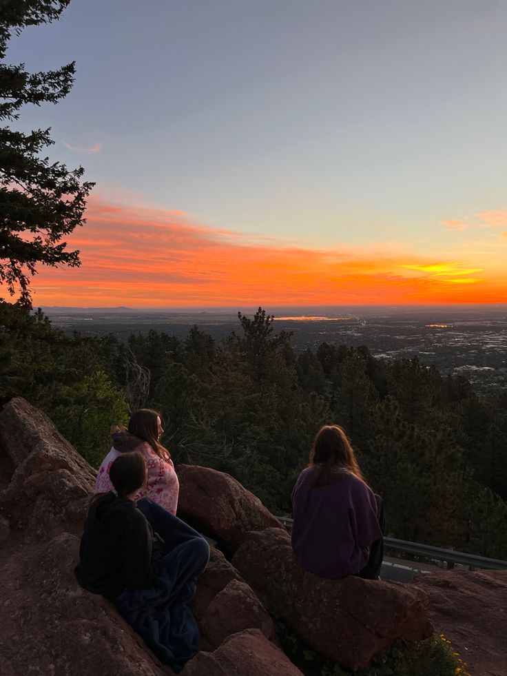 three people sitting on rocks looking at the sunset over a valley and city in the distance