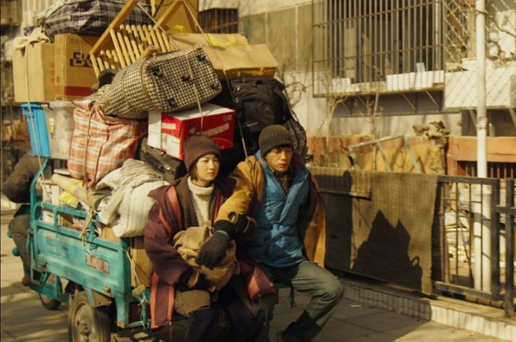 two people sitting on the back of a truck loaded with boxes and other items in front of a building