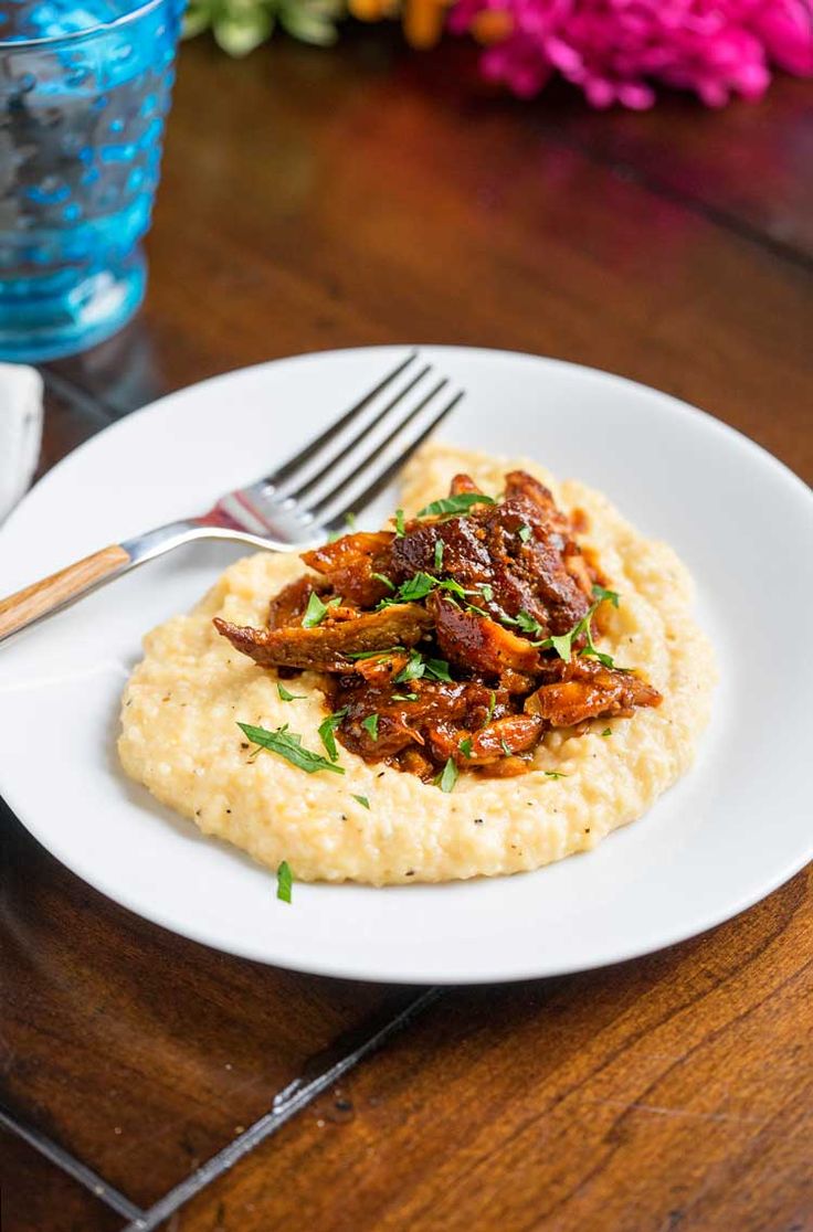 a white plate topped with meat and grits next to a fork on a wooden table