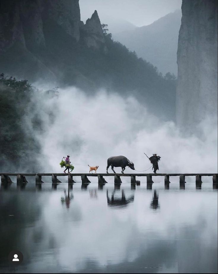 three people walking across a bridge over water with animals on the other side and mountains in the background