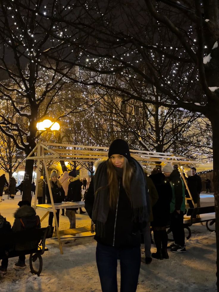 a woman walking in the snow at night with lights on trees and benches behind her