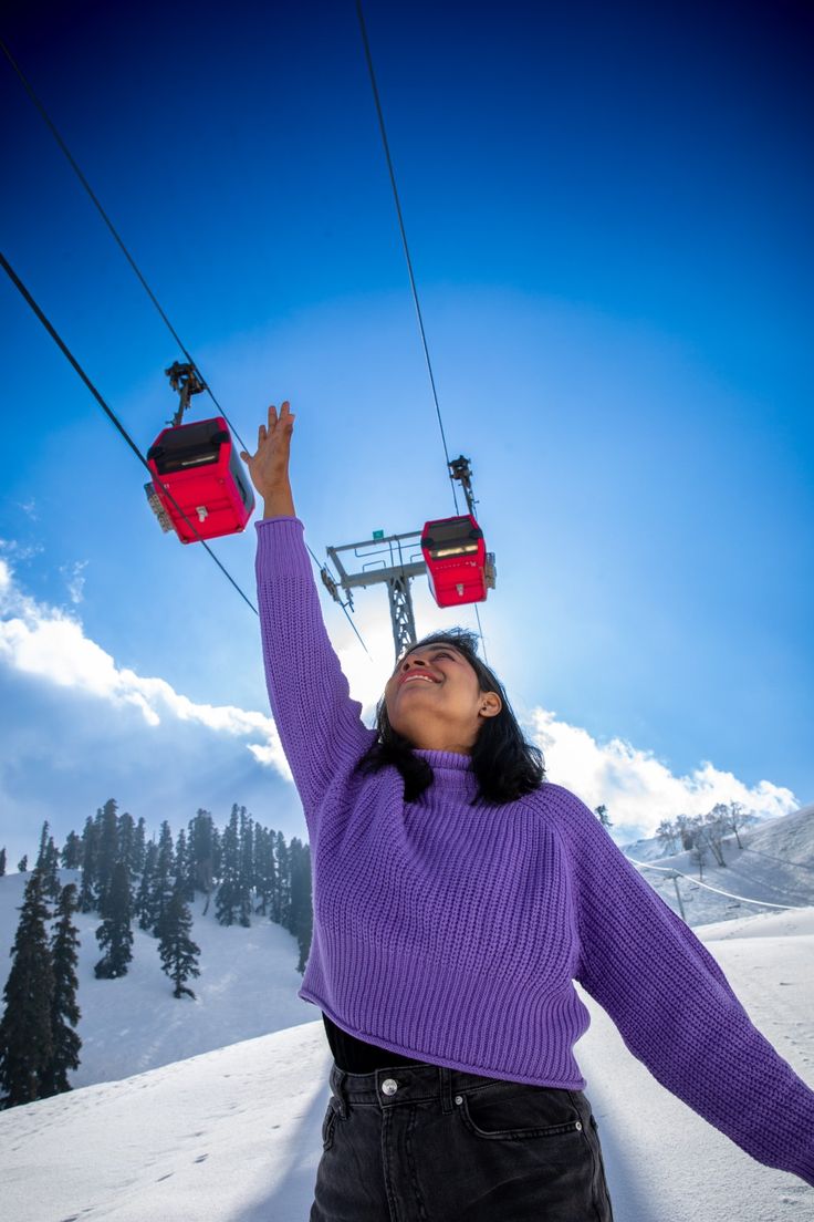 a woman in purple sweater standing on ski lift with her arms raised up to the sky