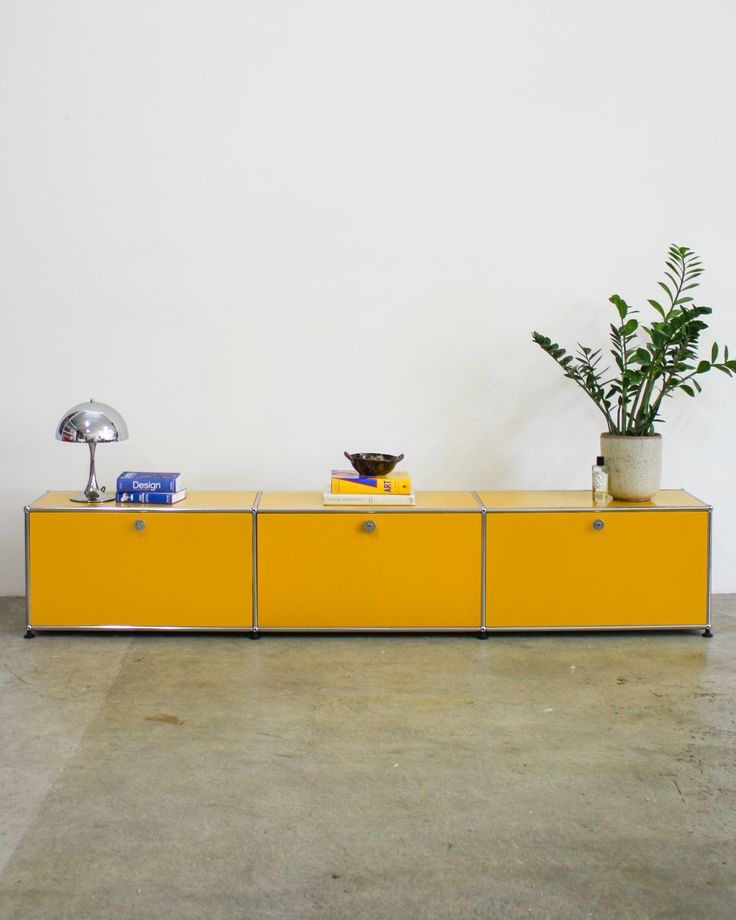 a yellow sideboard with two plants on top and a lamp next to it in front of a white wall