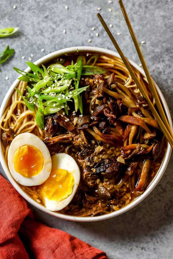 a bowl filled with noodles, meat and eggs on top of a gray surface next to chopsticks