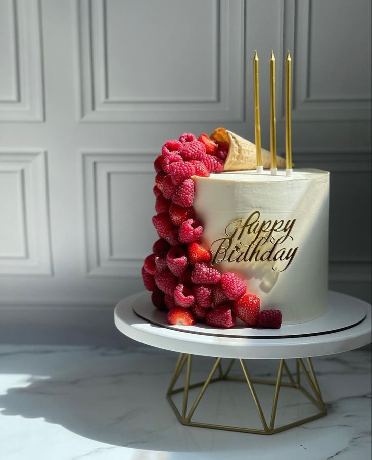 a birthday cake decorated with strawberries and gold candles sits on a white stand in front of a wall