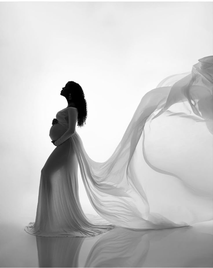 a pregnant woman in a white dress with flowing fabric around her neck and back, looking down at the ground