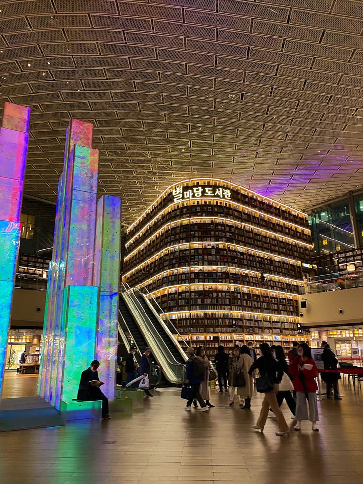 people are walking around in the lobby of an indoor shopping mall with brightly colored lights on the ceiling